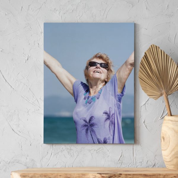 A memorial photo printed on a canvas, mounted on a wall. The photo shows an older woman grinning with her hands in the air next to the ocean.
