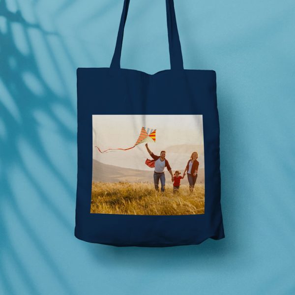 An image of a tote bag with a photo printed on the side of it. The photo is of a family running and flying a kite.